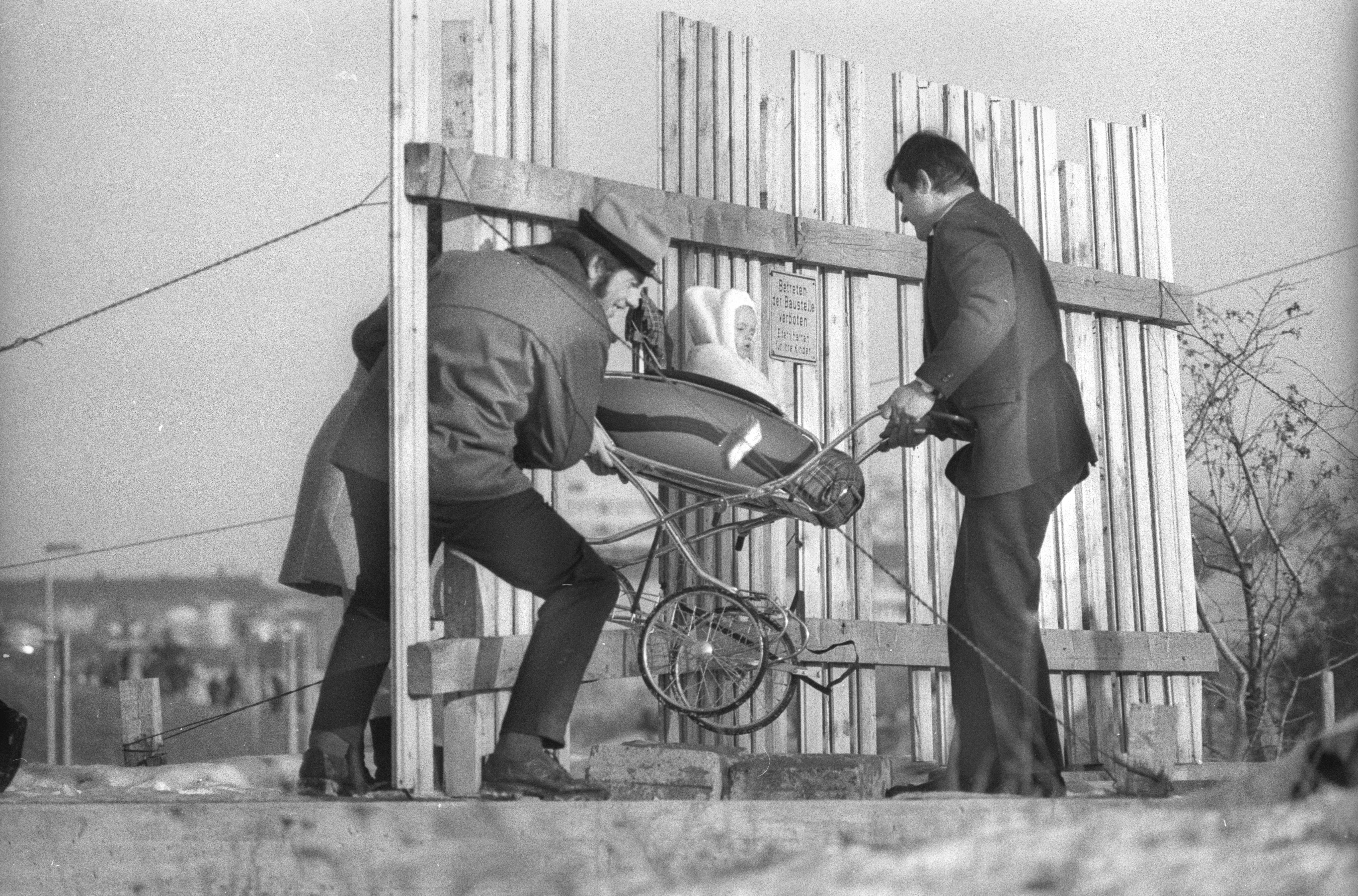 Baustelle auf dem Olympiagelände, 1970. Ein Kinderwagen wird durch den Zaun mit Schild "Betreten verboten" gehoben.
