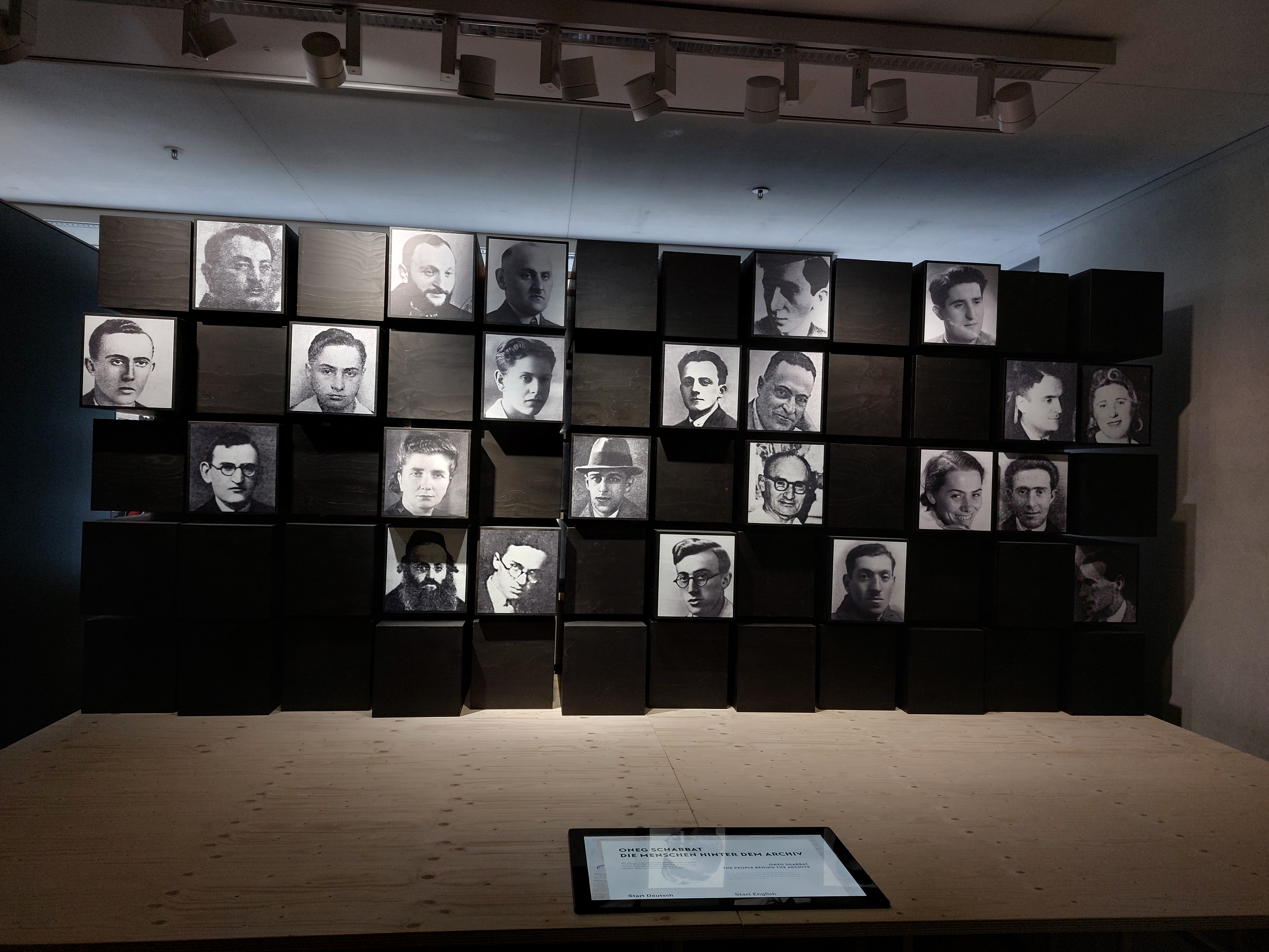 Fotowand mit Bildern der Gruppe Oneg Shabbat bestehend aus Frauen- und Männerportraits. Dazwischen sind schwarze leere Kästen, die die unbekannten Mitglieder symbolisieren.