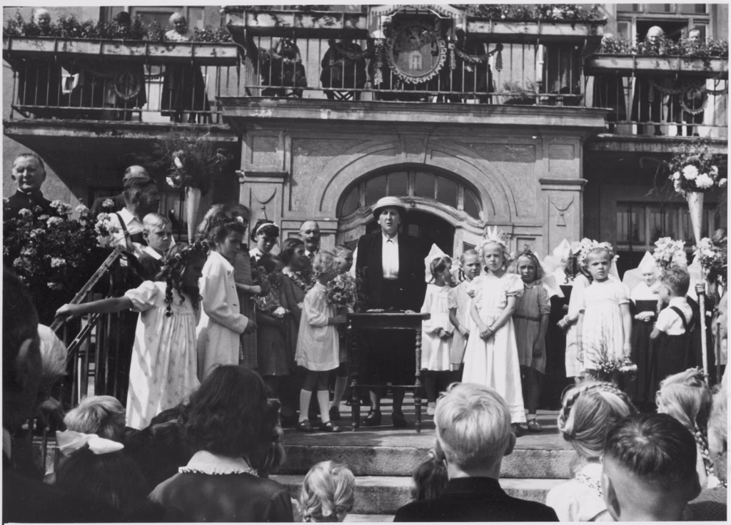 Das Bild zeigt Centa Aschenbrenner an einem Rednerpult bei einer Rede zu einem Fest vor dem Altenheim Heilggeist am Dom-Pedro-Platz umringt von Mädchen in weißen Kleidern, Nonnen und Geistlichen. Auf dem Balkon des Gebäudes hinter der Rednerin stehen Seniorinnen des Altenheims.